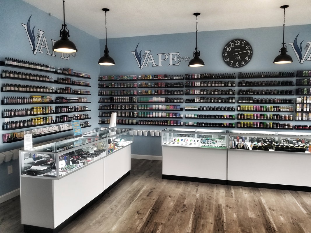 Interior Of A Vape Shop Featuring Shelves Filled With Various Vaping Products, Glass Display Cases, And A Wall Clock On A Light Blue Wall.